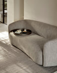 An Ethnicraft Ellipse Sofa - Ash sits on a matching rug in a minimalist room. On the sofa, a round black tray holds a book, a glass of water, and a small bowl. Soft natural light enters from the left.