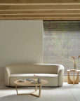 A minimalist living room features the Ethnicraft Ellipse Sofa in Oatmeal, a light wooden coffee table with ceramics, a side table with a vase, neutral walls, wooden ceiling beams, and sunlight streaming through window blinds.
