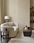 A minimalist living room features the Ethnicraft Ellipse Sofa in Oatmeal, a dark wood side table, and a brown coffee table with a lit candle and Jacques Deneef decor. Pottery-filled shelves and a lamp rest against neutral walls on a light rug.