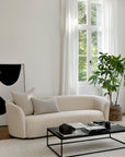 A modern living room with an Ethnicraft Ellipse Sofa in Oatmeal, a black-framed coffee table, a potted plant, a large window with white curtains, and abstract black-and-white artwork by Jacques Deneef leaning against the wall.