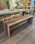 The SOBU Emily Bench - Walnut, with its natural finish, showcases stunning wood grain beside a long dining table on a rustic floor in a cozy, modern dining area.