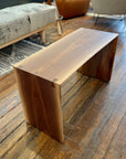 The SOBU Emily Bench - Walnut features a small, minimalist live edge design with clean lines and visible dovetail joints. Its natural walnut grain stands out on a rustic wooden floor among modern furniture.