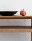 A black shallow bowl and a red pomegranate sit on the Emily Console - Oak by SOBU, set against a white textured wall.