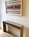 The Emily Console - Oak by SOBU sits against a beige wall beneath a colorful, geometric framed artwork with red stars, stripes, and patterns.