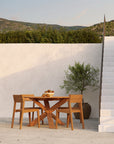 A teak outdoor dining table with three Ethnicraft EX1 Outdoor Dining Chairs sits on a patio by a white wall and staircase, topped with a potted plant and bowl of fruit. Green hills are visible in the background.