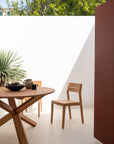 A minimalist outdoor patio with a round wooden table and two Ethnicraft EX1 Outdoor Dining Chairs in teak, complemented by a plant and ceramic pitcher, set against a white wall with natural sunlight and greenery.