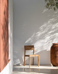 The Ethnicraft EX1 Outdoor Dining Chair in teak is placed on a sunlit patio beside a large terracotta pot, with leafy shadows softly cast over the white wall and floor.