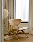 The Ethnicraft Eye Lounge Chair in oak is placed on a woven rug next to stacked books and a paper lantern, framed by tall beige curtains and a sheer white window.