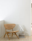 A minimalist room featuring the Ethnicraft Eye Lounge Chair - Oak next to a stack of books, a white modern lamp, and a beige cup, all set against a plain white wall and light European oak flooring.