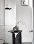A minimalist interior featuring Ethnicrafts Fin Side Table with sculptural legs, a metallic vase with bare branches, a chrome floor lamp, and abstract black-and-white art on a white wall.