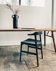 A minimalist room with a solid American ash table from the Frank Collection and a SOBU Frank Dining Chair in Black Ash. A tall black vase with red branches and a small white bowl adorn the table. Natural light streams in above; the floor is rustic wood.