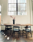 A minimalist dining area features a solid American ash table, three SOBU Frank Dining Chairs in black ash, and a black vase with red branches. Large windows fill the room with natural light, casting soft shadows on the wooden floor.