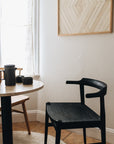 The Frank Dining Chair - Black Ash by SOBU sits beside a round table with cups and a teapot near a window with sheer curtains. Above the chair, geometric wooden art on the white wall evokes mid-century style.