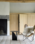 Minimalist living space with rustic solid oak floors, light wood cabinets, a black fireplace, the Ethnicraft Geometric Side Table in black oak, a butterfly chair with a fluffy fur throw, and an exposed wooden ceiling beam.