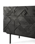 A close-up of the Ethnicraft Graphic Dresser, a modern black wooden cabinet with three drawers, crafted from solid teak and featuring intricate geometric carvings and slender legs.