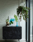A modern interior showcases the Ethnicraft Graphic Dresser in solid teak with a black geometric pattern, decorative vases with green leaves, a blue art print, stacked books, and a leafy hanging plant by a sunlit window.