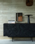 The Ethnicraft Graphic Sideboard - 3 door, featuring sleek black metal legs, stands against a concrete wall and is styled with art books, a black sculptural vase, and small artworks displayed above.