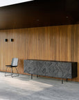 A modern minimalist room with vertical wooden slats, a Graphic Sideboard - 4 door by Ethnicraft in solid black teak, and a simple black chair on a gray floor. Two small wall lights are mounted above the sideboard.