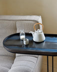 The Ethnicraft Graphite Organic Glass Tray - Oblong / Medium features a hand painted blue and black glass surface, displayed with a white cane-handled teapot, small cup, and sugar bottle beside a beige upholstered sofa.