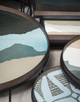 A set of Ethnicraft Graphite Wabi Sabi Glass Trays—round, extra-large tables with metal frames and abstract, layered blue, beige, white, and green tabletops evoking minimalist landscape art.