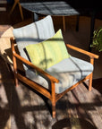 A wooden armchair with gray cushions and a SOBU Alma de Oaxaca Cushion - Butterfly sits in sunlight on a wooden floor, with window pane shadows cast across it and surrounding furniture in the background.