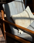 A SOBU wooden armchair with the Alma de Oaxaca Cushion in Fog sits in sunlight, its mid-century modern design casting shadows on a dark wooden floor. The chair features grey-blue linen cushions and a matching pillow.