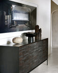 The Ethnicraft Grooves Sideboard - 3 door with a textured solid black stained teak finish stands against a white wall, adorned with decorative objects and topped by a large framed photograph of an industrial scene.