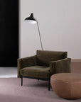 A modern lounge area with the Wooden Stories Attendant Lounge Chair in green velvet, French seams, paired with a round tan ottoman and a black floor lamp with a white inner shade, set against a minimalist backdrop.
