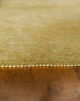 Close-up of the edge of the Radhi Rug - Moss by HIMALAYAN HERMITAGE, showing textured green Nepali wool and white stitching detail on a brown wooden floor, softly focused to highlight material and craftsmanship.