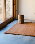 A small wooden stump sits on the Radhi Rug - Umber by HIMALAYAN HERMITAGE in a minimalist room with concrete floors and large windows letting in natural light.