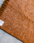 A close-up of the Radhi Rug - Umber by HIMALAYAN HERMITAGE, a rust-colored handwoven wool rug with fringe, displayed on a gray concrete floor. A white fabric label near the fringe highlights its custom craftsmanship.