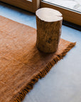 The Radhi Rug - Umber by HIMALAYAN HERMITAGE, with fringed edges, lies on a smooth gray floor next to a large window, topped by a wooden stump stool.