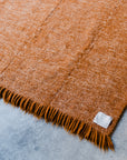 A close-up of the HIMALAYAN HERMITAGE Radhi Rug - Umber, a rust-colored handwoven wool rug with fringed edges and a small white label, displayed on a light gray concrete floor.