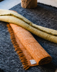 Two rolled-up rugs, one orange with fringes and one light green, rest atop the HIMALAYAN HERMITAGE Radhi Rug - Ink, a large dark gray handwoven wool rug, placed on a smooth floor near a window.