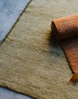 A close-up of the HIMALAYAN HERMITAGE Radhi Rug - Moss on a gray concrete floor, featuring a white label in the corner and an orange-brown fringed blanket partially rolled up on the rug.
