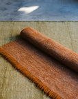 The HIMALAYAN HERMITAGE Radhi Rug - Moss, with a brown textured hand-woven design, is partially rolled out over a larger green rug on a gray floor, bathed in sunlight.