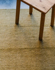 A close-up of a wooden table resting on the HIMALAYAN HERMITAGE Radhi Rug - Moss, a textured, light green handwoven rug. Natural light highlights the visible wood grain and artisanal weave.