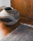 A large, rustic clay pot sits on a wooden floor next to the HIMALAYAN HERMITAGE Radhi Rug - Silver with fringed edges and a wooden cabinet in a cozy, sunlit room.
