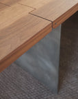 Close-up of the SOBU Hinge Extension Dining Table - Black Walnut, showcasing its zigzag interlocking joint detail and gray metal leg, set on a textured carpeted floor.