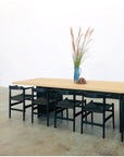 The SOBU Hinge Extension Dining Table in White Ash, paired with six black chairs, is set against a plain white wall. On the table are a blue vase with tall grass and a small centerpiece, atop a concrete floor.