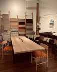A modern showroom displays the SOBU Hinge Extension Dining Table in white oak with four orange-woven chairs, rug wall displays, shelves, a couch with patterned pillows, and wall art in the background.