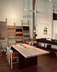 A person with long hair sits at the SOBU Hinge Extension Dining Table - White Oak in a modern, cozy room with woven rugs on ladders, bench cushions, and a decorative airplane model hanging on the wall.