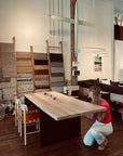 A woman in a red skirt and white shirt crouches next to a SOBU Hinge Extension Dining Table - White Oak in a modern showroom featuring textile wall displays, chairs, and hanging art pieces.