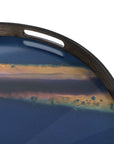A close-up of the Ethnicraft Indigo Organic Glass Tray (Round / Small), showcasing organic forms on a blue surface with gold and brown streaks resembling a horizon, and an oval cut-out handle near the edge.