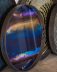 The Indigo Organic Glass Tray - Round / Small by Ethnicraft features a vibrant blue, purple, orange, and gold abstract design with organic forms and sits upright on a wooden shelf among other patterned trays.