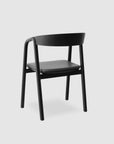 The Wooden Stories Inlay Dining Chair in black oak and leather features a curved plywood backrest, four straight legs, and a flat seat, offering a sleek, minimalist look against a plain light gray background.