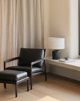 A minimalist room with Scandinavian modernism, featuring the Ethnicraft Jack Footstool in black leather beside a matching armchair on light wood floors, beige curtains, and a white wall shelf holding a lamp, cup, and book near the window.