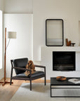 A brown dog sits on an Ethnicraft Jack Lounge Chair in black leather in a modern living room with a white wall, fireplace, floor lamp, minimalist decor, and a low black coffee table inspired by Scandinavian modernism.
