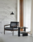 Modern minimalist interior with the Jack Lounge Chair - Black Leather by Ethnicraft, a matching side table, books, glass, floor lamp, and large window with sheer curtains—an inviting space inspired by Scandinavian modernism.