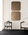 A minimal interior with two square bronze mirrors, a round side table with a white vase and book, and an Ethnicraft Jack Lounge Chair - Ivory featuring dark wooden arms and legs, on a dark floor beside sheer curtains.